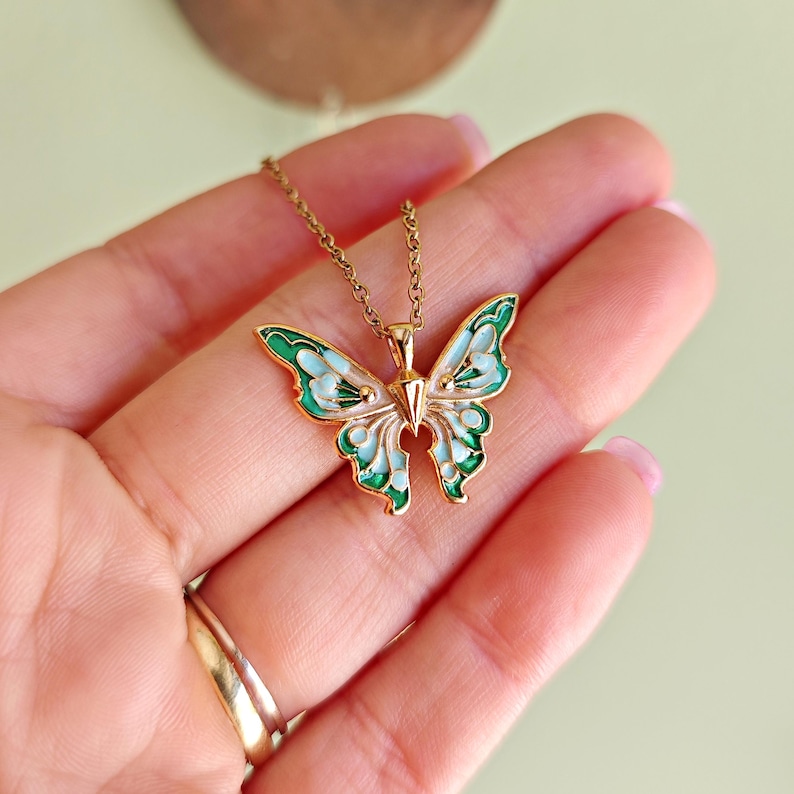 May include: A gold-colored butterfly pendant necklace. The butterfly wings are detailed with green and light blue enamel, and the necklace chain is delicate. The pendant is held in a hand, showcasing the jewelry's design and color.