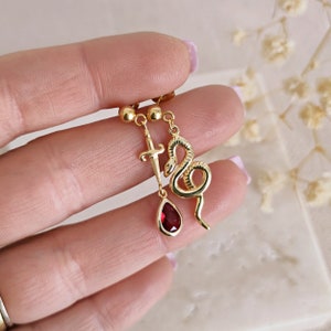 May include: Gold dangle earrings with a red gemstone, a snake charm, and a sword charm.