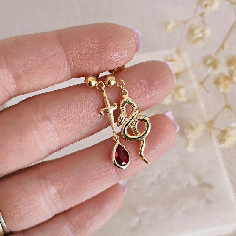May include: Gold dangle earrings with a red gemstone, a snake charm, and a sword charm.