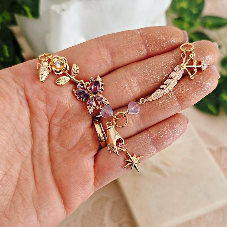 May include: A gold chain bracelet with charms including a rose, butterfly, hand, star, and feather. The charms are decorated with purple and clear crystals.