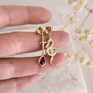 May include: Gold earrings with a dangling red gemstone, a snake charm, and a dagger charm.