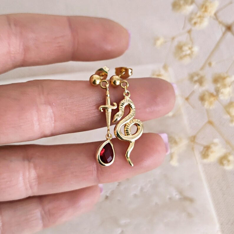 May include: Gold earrings with a dangling red gemstone, a snake charm, and a dagger charm.