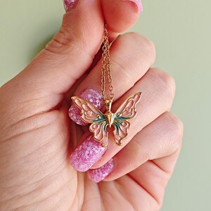 May include: Gold-toned butterfly pendant necklace with a delicate chain. The butterfly has a detailed design with pink and teal enamel accents. The necklace is held against a background of a hand with pink nail polish.