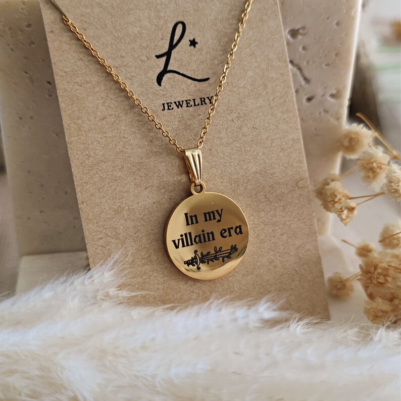 May include: Gold necklace with a round pendant that says "In my villain era" with a small sword design. The necklace is on a brown card with the text "L* Jewelry".