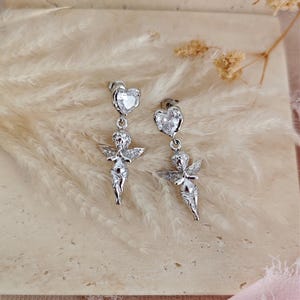 May include: A pair of silver-colored dangle earrings. Each earring features a heart-shaped stud with a clear stone, connected to a small cherub figure with outstretched wings. The earrings are displayed against a soft, neutral background.