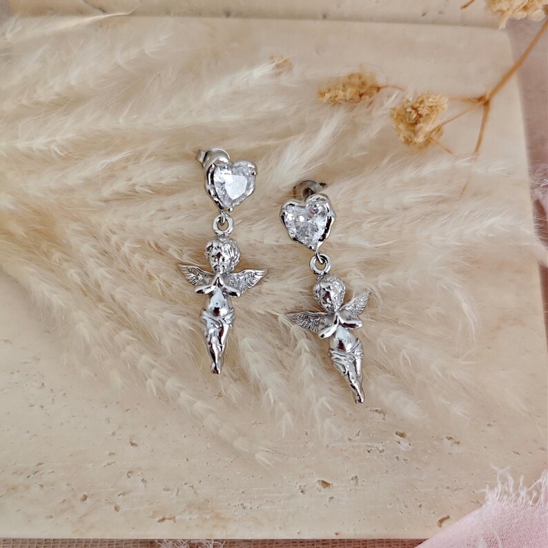 May include: A pair of silver-colored dangle earrings. Each earring features a heart-shaped stud with a clear stone, connected to a small cherub figure with outstretched wings. The earrings are displayed against a soft, neutral background.