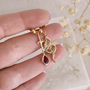 May include: Gold dangle earrings with a red gemstone, a snake, and a sword charm.