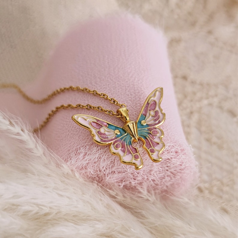 May include: A gold-colored butterfly pendant necklace with a delicate chain. The butterfly has a detailed design with pink, teal, and white enamel. The necklace is displayed on a soft pink surface.