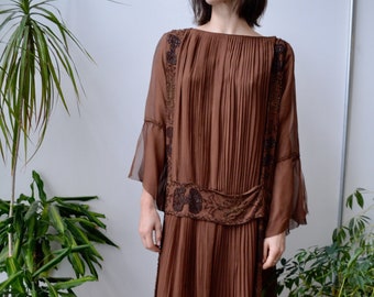 May include: A brown pleated dress with beaded embellishments and sheer sleeves. The dress is knee-length and has a relaxed fit. The model is wearing white platform sandals.