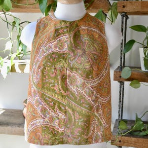 May include: A sleeveless top with a paisley pattern in shades of olive green, orange, and pink. The top is displayed on a mannequin and features a round neckline and a button closure. The fabric has a vintage aesthetic.