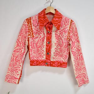 Puede incluir: Una chaqueta corta de estampado rosa y blanco con cierre de botones y mangas largas. La chaqueta tiene un diseño de paisley.