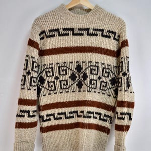 Puede incluir: Suéter beige con patrones geométricos marrones y negros. El suéter tiene cuello redondo y mangas largas. El diseño incluye rayas horizontales y formas geométricas repetidas. El suéter está colgado en una percha de madera.