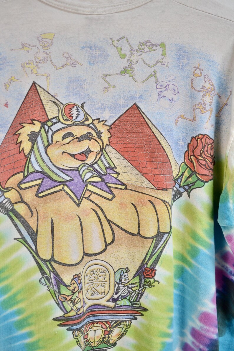 May include: A light-colored t-shirt featuring a colorful graphic of a bear wearing a headdress, pyramids, and skeletons. The bear holds a rose. The shirt has a tie-dye pattern on the sleeve.