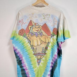 May include: Tie-dye t-shirt featuring a colorful graphic of a bear-like character with roses, pyramids, and a decorative base. The shirt has a rainbow tie-dye pattern in shades of blue, green, purple, and yellow. The top of the shirt has a light beige color with skeleton figures.