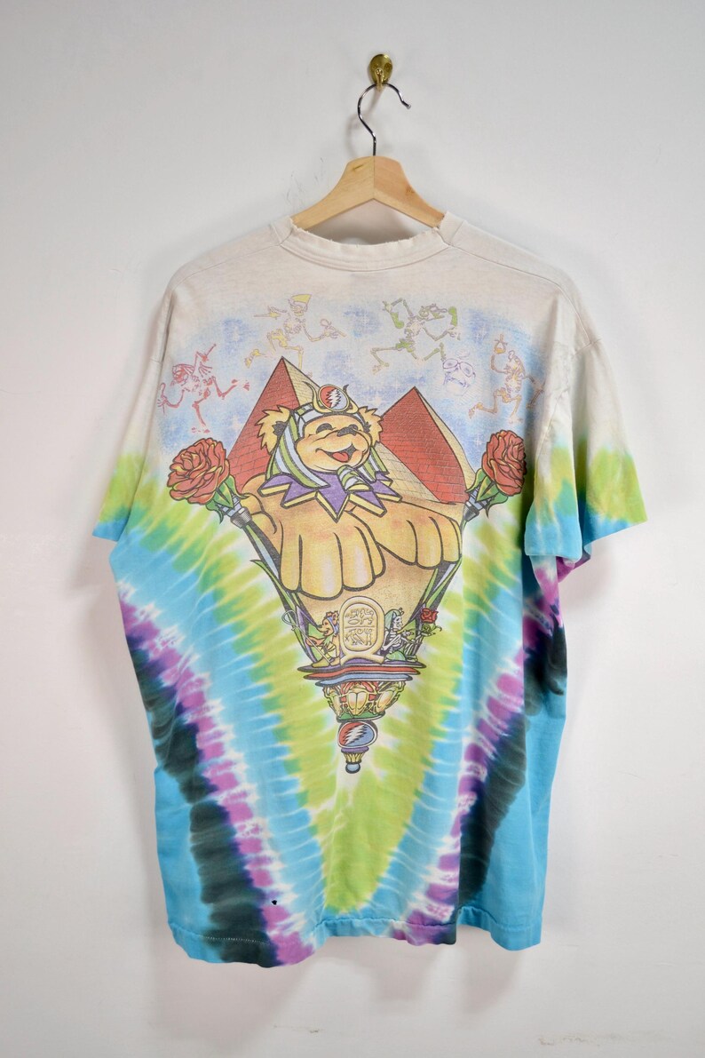 May include: Tie-dye t-shirt featuring a colorful graphic of a bear-like character with roses, pyramids, and a decorative base. The shirt has a rainbow tie-dye pattern in shades of blue, green, purple, and yellow. The top of the shirt has a light beige color with skeleton figures.