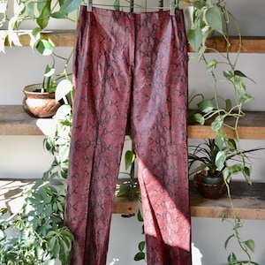 May include: Red faux leather trousers with a snakeskin pattern hanging on a wooden hanger. The trousers are straight-legged and have a high waist. The background includes wooden shelves with plants.