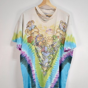 May include: Vintage tie-dye t-shirt with a colorful graphic. The shirt features a central design of cartoon characters in a decorative chalice, surrounded by a spiral tie-dye pattern in blue, green, purple, and yellow.
