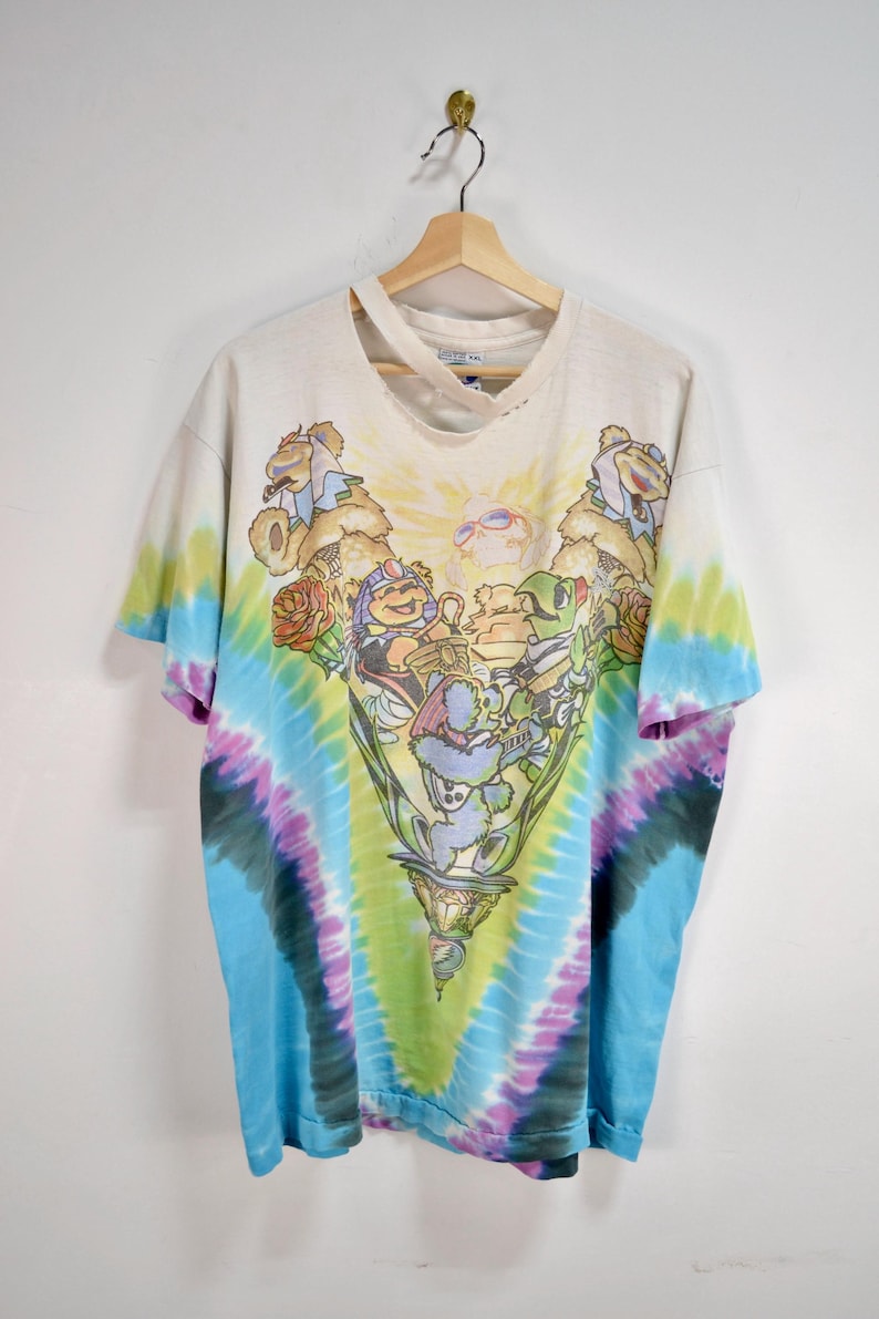 May include: Vintage tie-dye t-shirt with a colorful graphic. The shirt features a central design of cartoon characters in a decorative chalice, surrounded by a spiral tie-dye pattern in blue, green, purple, and yellow.
