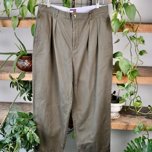 May include: Olive green pleated trousers hanging on a wooden hanger. The trousers have a button closure and a small logo detail. The trousers are displayed against a backdrop of wooden shelves and greenery.