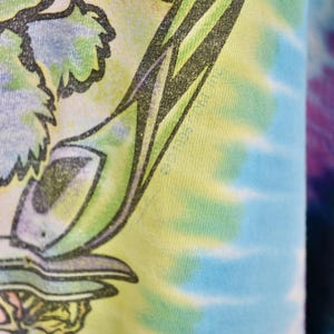 May include: Close-up of a tie-dye t-shirt with a colorful design. The shirt features a graphic with green, blue, and yellow hues. The tie-dye pattern is visible on the right side of the image.