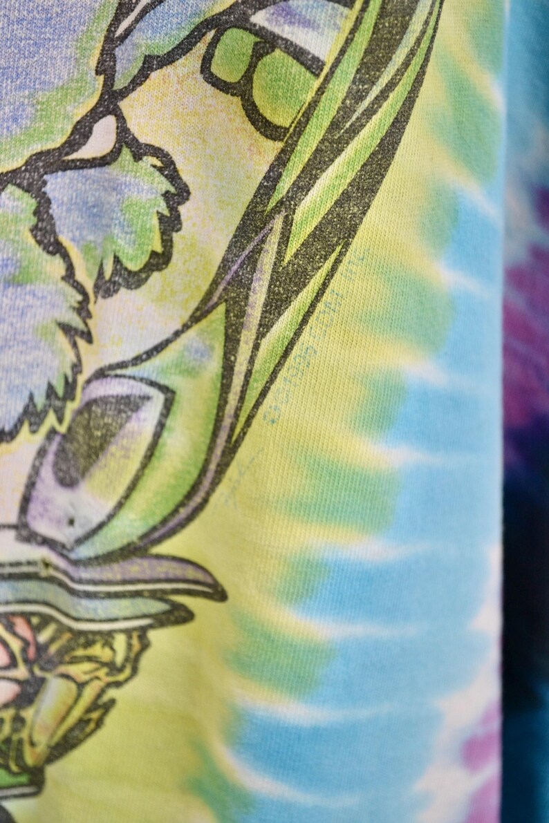 May include: Close-up of a tie-dye t-shirt with a colorful design. The shirt features a graphic with green, blue, and yellow hues. The tie-dye pattern is visible on the right side of the image.