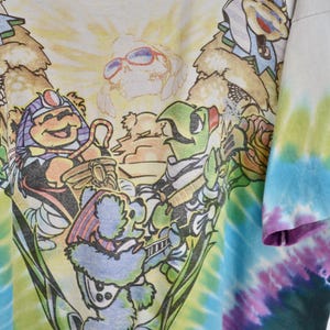 May include: White t-shirt with a colorful graphic of cartoon bears playing musical instruments. The design is set against a tie-dye background of blue, green, and purple hues. The shirt has a distressed neckline.