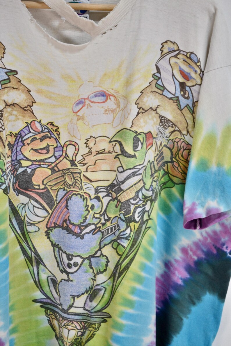 May include: White t-shirt with a colorful graphic of cartoon bears playing musical instruments. The design is set against a tie-dye background of blue, green, and purple hues. The shirt has a distressed neckline.