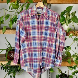 May include: A long-sleeved plaid shirt in shades of red, blue, and yellow hangs on a wooden hanger. The shirt has a classic collar and button-down front. The background includes wooden shelves and green plants.