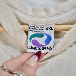 May include: Close-up of a cream-colored t-shirt tag with the text "100% COTTON MADE IN USA XXL CARE ON REVERSE". The tag features a Liquid Blue logo with green, purple, and blue colors.