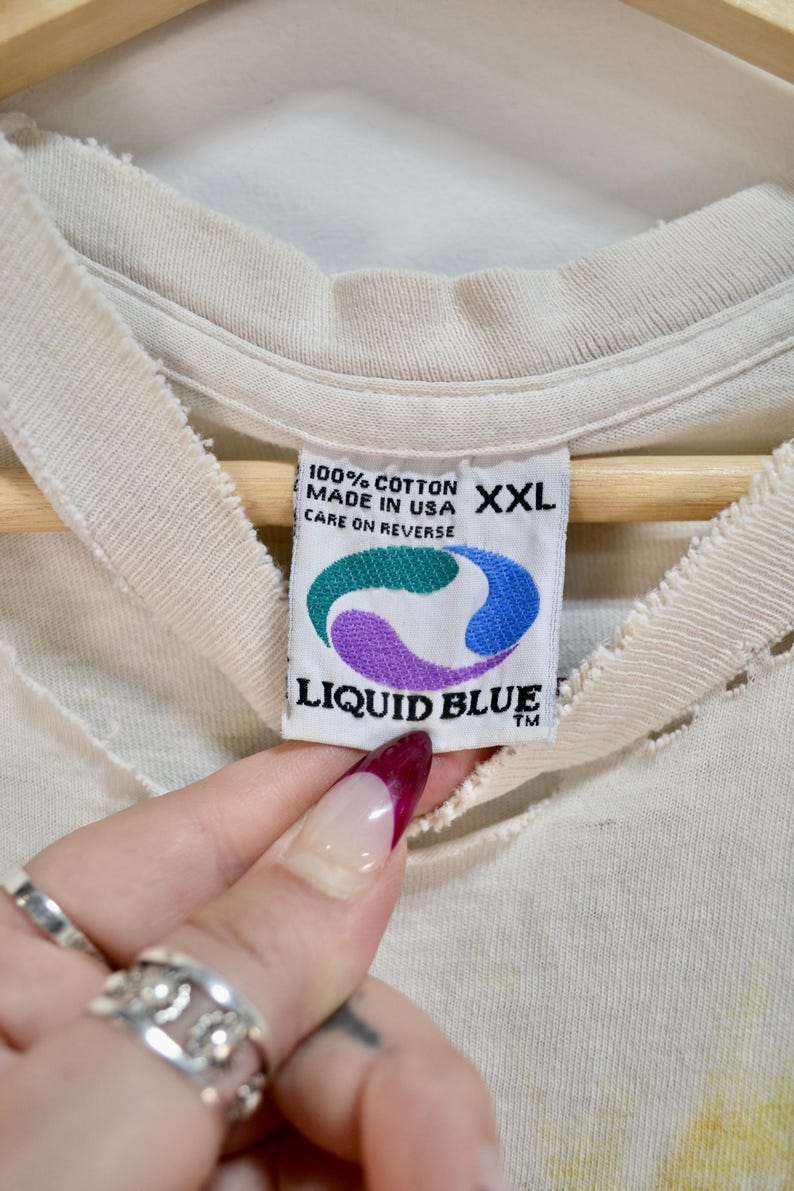 May include: Close-up of a cream-colored t-shirt tag with the text "100% COTTON MADE IN USA XXL CARE ON REVERSE". The tag features a Liquid Blue logo with green, purple, and blue colors.