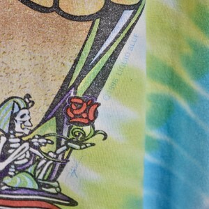 May include: Close-up of a tie-dye t-shirt featuring a skeleton holding a rose. The skeleton is wearing a hat and is surrounded by green, purple, and red colors. The shirt has a colorful tie-dye pattern in shades of blue, green, and yellow.