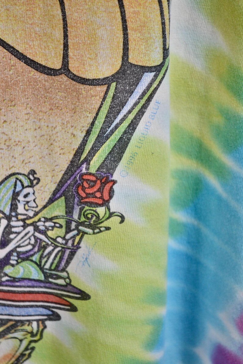 May include: Close-up of a tie-dye t-shirt featuring a skeleton holding a rose. The skeleton is wearing a hat and is surrounded by green, purple, and red colors. The shirt has a colorful tie-dye pattern in shades of blue, green, and yellow.