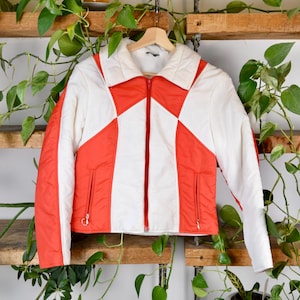 May include: A retro ski jacket in white and orange, featuring a full zip and two front pockets. The jacket has a geometric pattern with orange diamond shapes and white sections. It is displayed on a wooden hanger.