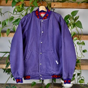 May include: A purple bomber jacket with a button-up front, two front pockets, and a red and black patterned collar and cuffs. The jacket is hanging on a wooden hanger against a backdrop of wooden shelves and green plants.