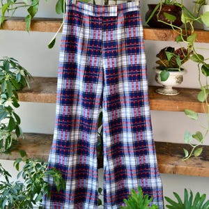 May include: Vintage plaid trousers in blue, white, and red. The high-waisted trousers are hanging on a wooden hanger. The trousers have a wide leg and a button closure. The background includes wooden shelves and green plants.