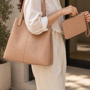 Puede incluir: Un bolso tote beige y un estuche rectangular con cremallera a juego, ambos con un patrón texturizado de imitación de cocodrilo. El bolso tiene dos correas para el hombro y el estuche incluye una correa de muñeca. La palabra Qoda es visible.