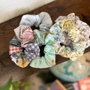 May include: A collection of colorful scrunchies in various floral and patterned fabrics. The scrunchies are in shades of blue, pink, yellow, and green. They are arranged on a dark wooden surface, showcasing their textures and designs.