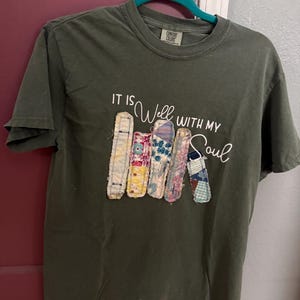 May include: Olive green t-shirt with the phrase "IT IS Well WITH MY Soul" in white script. The shirt features a design of colorful fabric strips. The shirt is hanging on a teal hanger.
