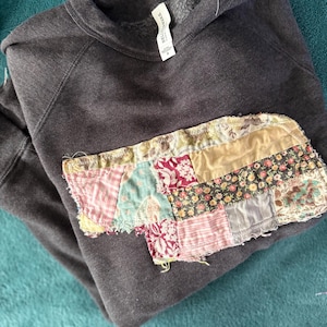 May include: Dark gray sweatshirt with a patchwork design on the front. The patchwork features various fabric patterns in shades of pink, green, yellow, and floral prints. The sweatshirt is folded and resting on a teal surface.