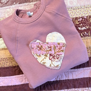 May include: A mauve-colored sweatshirt with a heart-shaped patch. The patch is made of floral fabric in shades of pink, cream, and burgundy. The sweatshirt is folded on a patterned quilt.