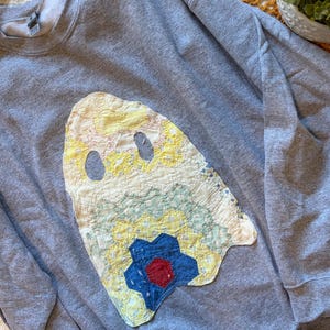 May include: A heather gray crewneck sweatshirt featuring a large, ghost-shaped appliqué made from vintage-style quilted fabric. The ghost design is constructed from various pastel-colored floral patches, including yellow, light blue, and a dark blue star.