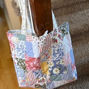 May include: A patchwork tote bag with a variety of floral, striped, and checkered fabric squares in shades of blue, red, yellow, and green. The bag has a quilted appearance and white straps.