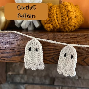 Könnte beinhalten: Eine Häkelgirlande mit weißen Geisterornamenten und schwarzen Augen. Die Girlande hängt an einer Holzoberfläche. Der Text "Crochet Pattern" ist auf einem braunen Rechteck geschrieben.