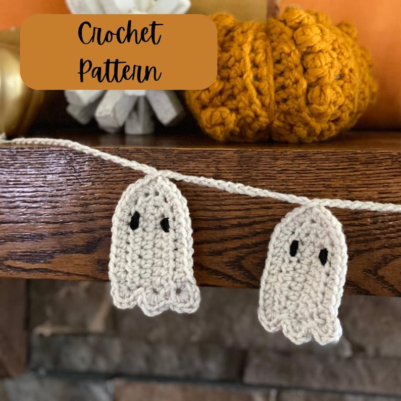 May include: A crochet garland with white ghost ornaments and black eyes. The garland is hanging on a wooden surface. The text "Crochet Pattern" is written on a brown rectangle.