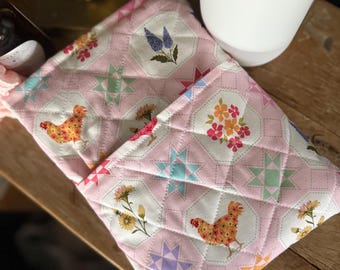 May include: Pink quilted pot holders with a repeating pattern of chickens, flowers, and starbursts. The pot holders are made of a soft fabric and have a diamond-stitched pattern. The design includes various colors such as yellow, orange, blue, and pink.