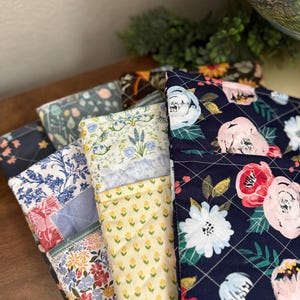 May include: A stack of quilted fabric pouches with floral patterns. The pouches feature a variety of designs, including blue and white flowers, yellow floral patterns, and a navy blue floral print. The fabrics are quilted with a diamond pattern.