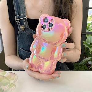 May include: A glossy, iridescent pink phone case shaped like a teddy bear. The case features a smooth, reflective surface with a pearlescent finish that shifts colors. It has precise cutouts for a triple-lens camera system on the back.