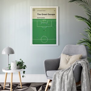 May include: A framed poster titled "The Great Escape" featuring a football play diagram. The poster has a white border and is displayed on a light blue wall. The room includes a gray armchair, a side table with a lamp, and a potted plant.