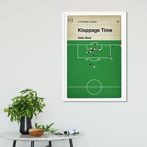 May include: A white framed poster with a green soccer field and a white circle in the center. The poster has the text "a Football Classic" and "Klopppage Time" in black text. The text "Sadio Man&eacute;" is also in black text. There are white and black circles on the field.