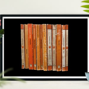 Puede incluir: Una impresión enmarcada de una colección de 15 libros de bolsillo de color naranja y marrón con el texto "Christopher Nolan" en las cubiertas. Los libros están dispuestos verticalmente sobre un fondo negro.